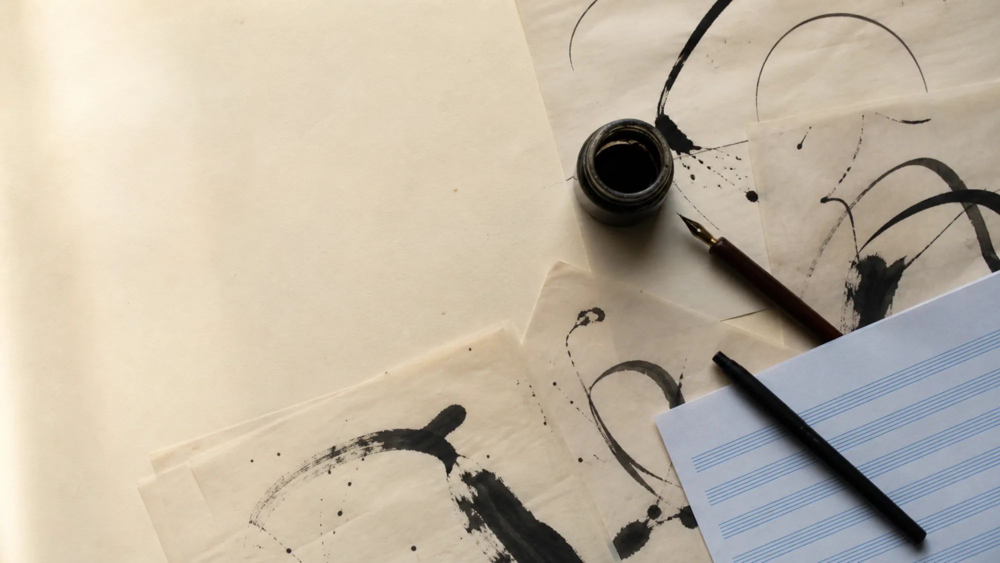An open ink bottle and dip pen resting on sheets of brush-lettering practice and blank manuscript paper.